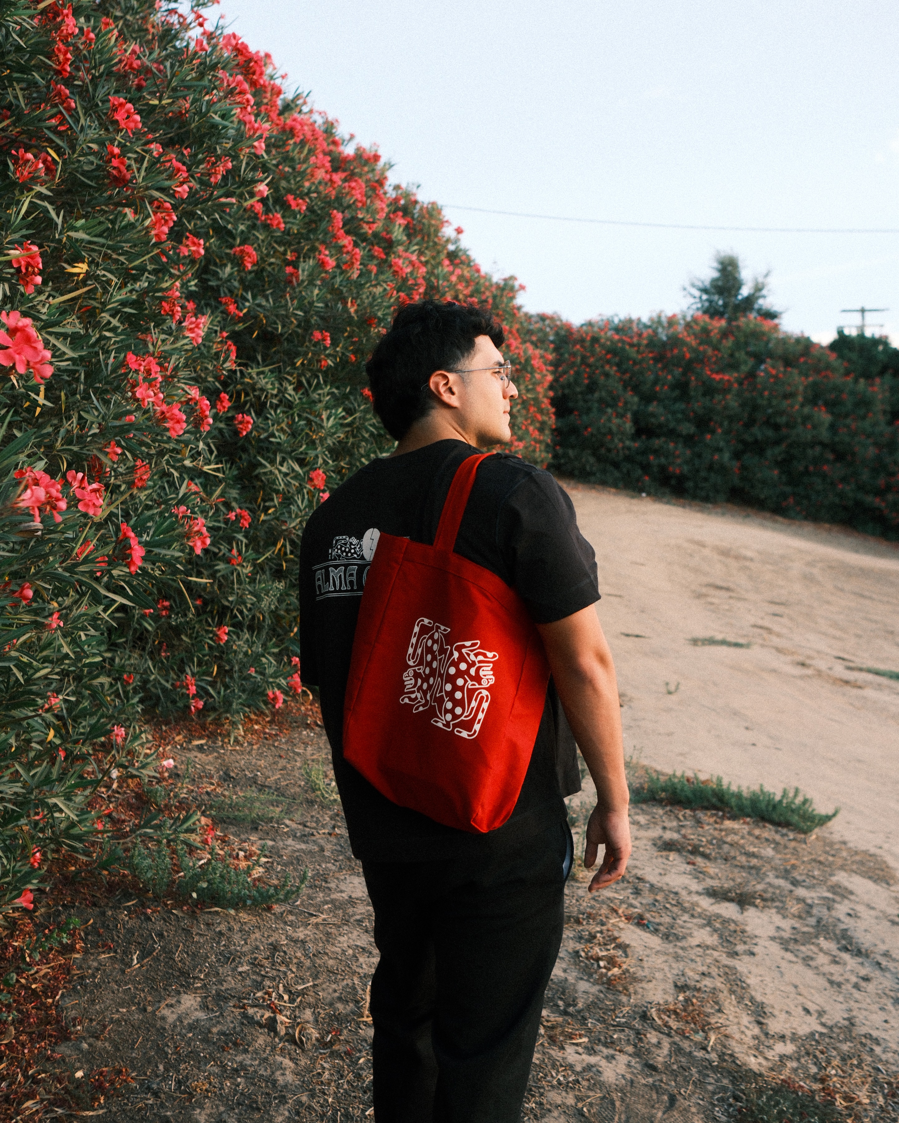 Alma Oscura — customer walking along a hedge of flowering oleander carrying the red Alma Oscura tote bag