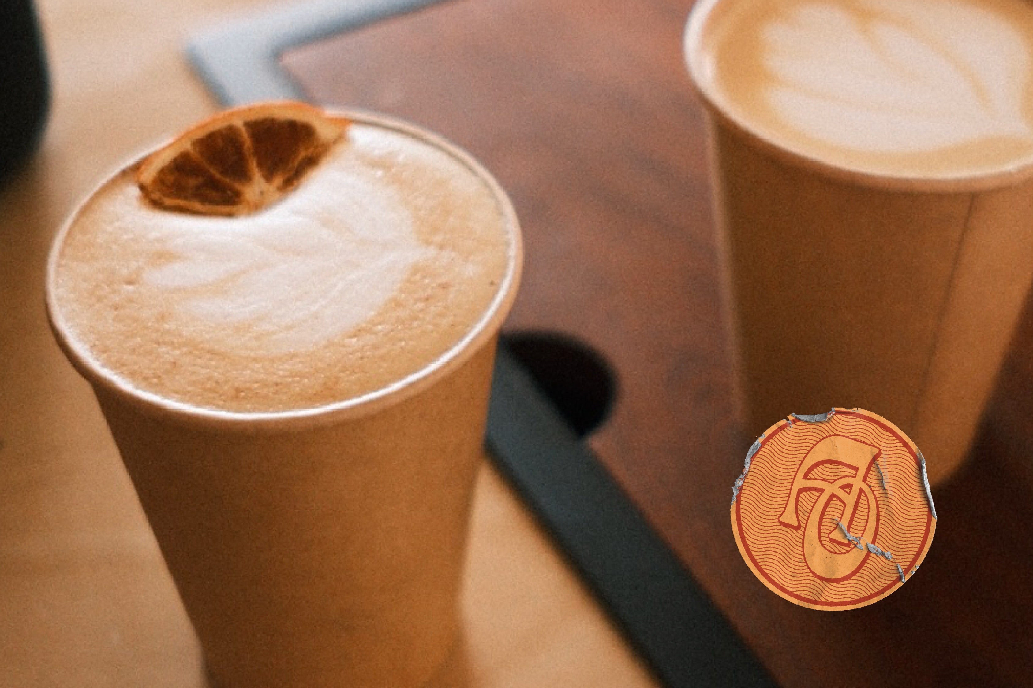 Alma Oscura — two lattes in kraft cups on the counter, one topped with a dried orange slice, with the AO monogram sticker