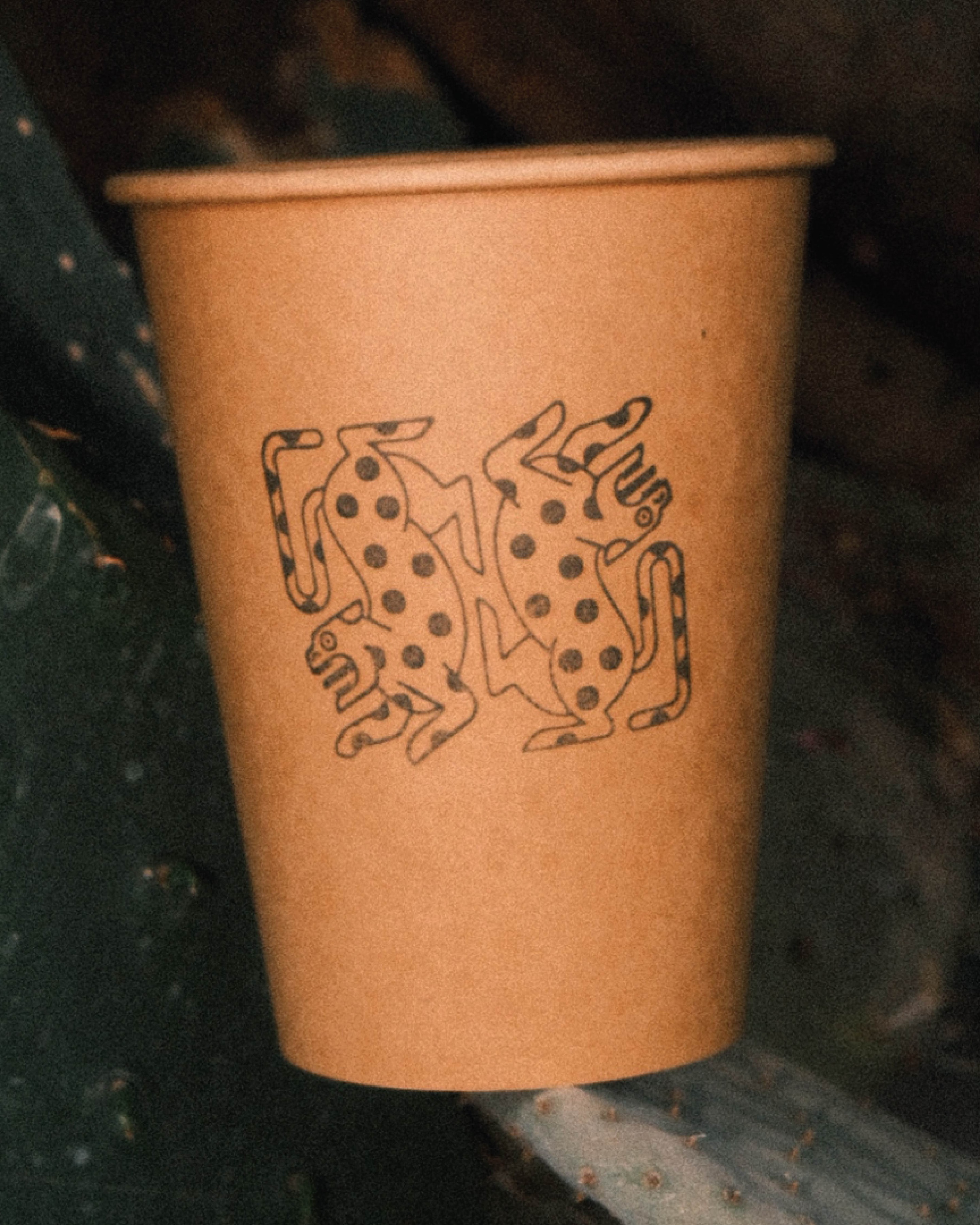 Alma Oscura — kraft paper cup printed with the twin jaguar illustration, resting on a prickly pear cactus