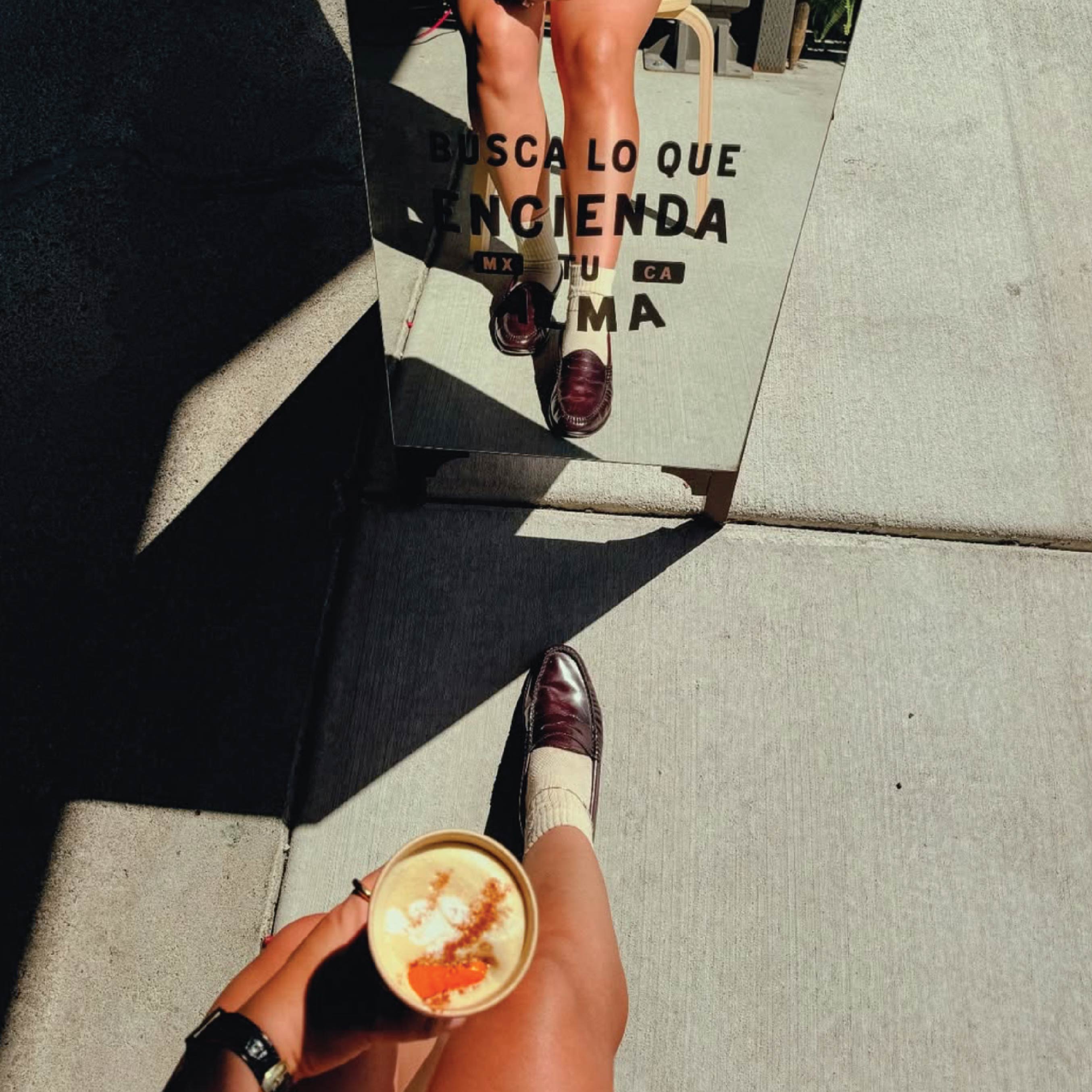 Alma Oscura — customer holding a latte beside the Busca Lo Que Encienda Tu Alma mirror sign in hard afternoon light